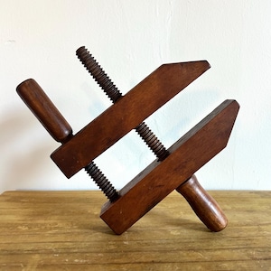 May include: A dark brown wooden hand screw clamp featuring two parallel jaws connected by two threaded wooden spindles with integrated handles, designed for securing materials together during woodworking or assembly tasks.