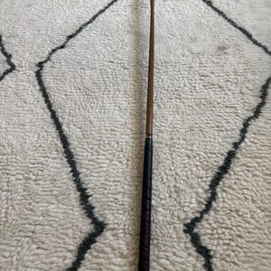 May include: A golf putter featuring a silver-toned metal head, a long wooden shaft, and a black textured grip. The club is positioned vertically, showcasing the contrast between the metallic head, the smooth wood, and the wrapped handle.