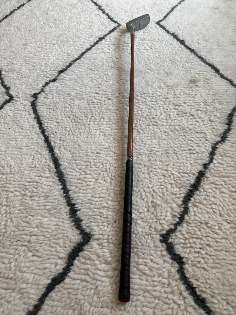 May include: A golf putter featuring a silver-toned metal head, a long wooden shaft, and a black textured grip. The club is positioned vertically, showcasing the contrast between the metallic head, the smooth wood, and the wrapped handle.