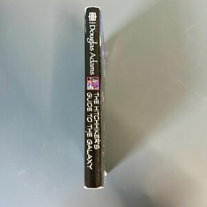 May include: A copy of "The Hitchhiker's Guide to the Galaxy" by Douglas Adams. The book's spine is black with white lettering. The cover features a colourful illustration. The word "Fiction" is at the top.