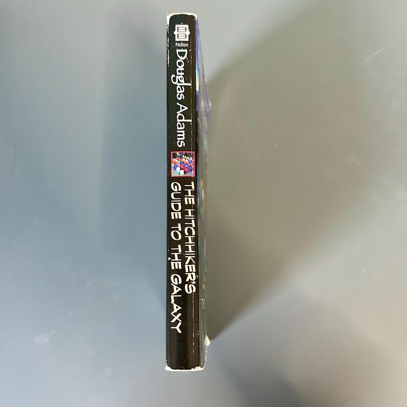 May include: A copy of "The Hitchhiker's Guide to the Galaxy" by Douglas Adams. The book's spine is black with white lettering. The cover features a colourful illustration. The word "Fiction" is at the top.