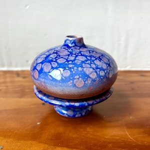 May include: A small ceramic bud vase with a rounded body and a pedestal base. The glaze features a vibrant blue color with crystalline patterns in shades of light purple and white, transitioning to a brown clay tone at the bottom.