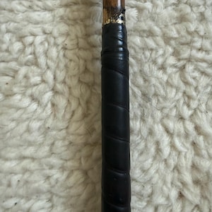 May include: A wooden golf club shaft featuring a black leather grip wrapped in a spiral pattern. The transition point between the wood and the grip shows a small section of gold-colored lettering on a black band, indicating the club's construction.