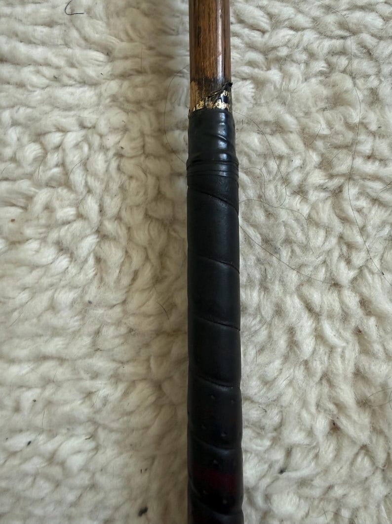 May include: A wooden golf club shaft featuring a black leather grip wrapped in a spiral pattern. The transition point between the wood and the grip shows a small section of gold-colored lettering on a black band, indicating the club's construction.