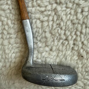 May include: A golf putter featuring a wooden shaft and a silver-toned metal head with a textured surface. The head displays a vertical line across the center and the engraved signature text "Oty Crisman" on the lower face of the club head.