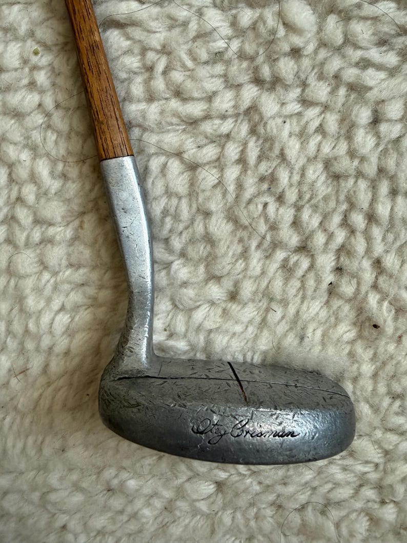 May include: A golf putter featuring a wooden shaft and a silver-toned metal head with a textured surface. The head displays a vertical line across the center and the engraved signature text "Oty Crisman" on the lower face of the club head.