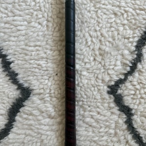 May include: A golf club grip featuring a dark, textured leather wrap with a spiral pattern. The grip transitions into a smooth, light brown wooden shaft at the top, secured by a metallic ferrule. The handle has a cylindrical shape and a dark finish.