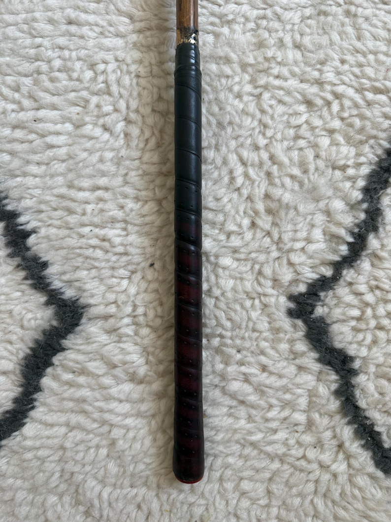 May include: A golf club grip featuring a dark, textured leather wrap with a spiral pattern. The grip transitions into a smooth, light brown wooden shaft at the top, secured by a metallic ferrule. The handle has a cylindrical shape and a dark finish.