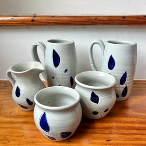 May include: A collection of five ceramic pieces featuring a light gray speckled glaze and dark blue leaf-shaped accents. The set includes two tall mugs with handles, a small pitcher with a spout, and two rounded bowls with wide openings.