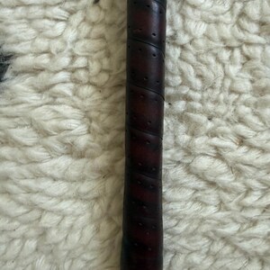 May include: A dark brown, cylindrical handle featuring a spiral-wrapped, perforated texture. The grip has a smooth, slightly worn appearance with visible stitching along the diagonal wrap, terminating in a small, rounded red base at the bottom.