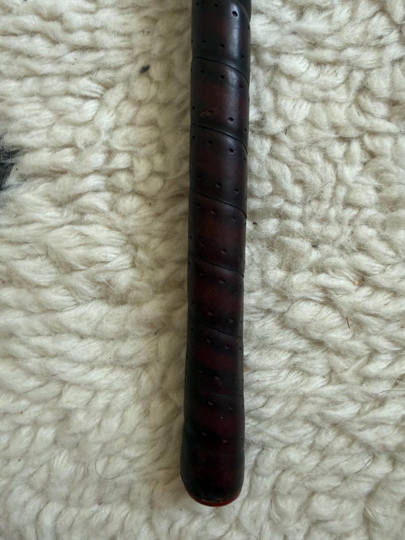May include: A dark brown, cylindrical handle featuring a spiral-wrapped, perforated texture. The grip has a smooth, slightly worn appearance with visible stitching along the diagonal wrap, terminating in a small, rounded red base at the bottom.