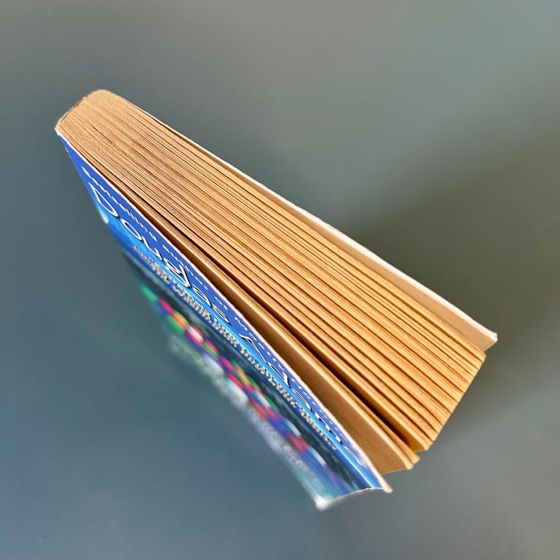 May include: A hardback book with a blue cover and gold-toned pages. The book is slightly open, revealing the pages and the title. The cover has colourful text and graphics. The book is angled, showing the side view of the pages.
