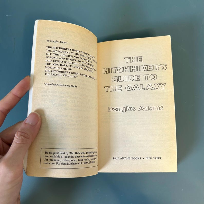 May include: An open book titled "The Hitchhiker's Guide to the Galaxy" by Douglas Adams. The book's pages are aged, with black text on a yellow background. Published by Ballantine Books, New York.