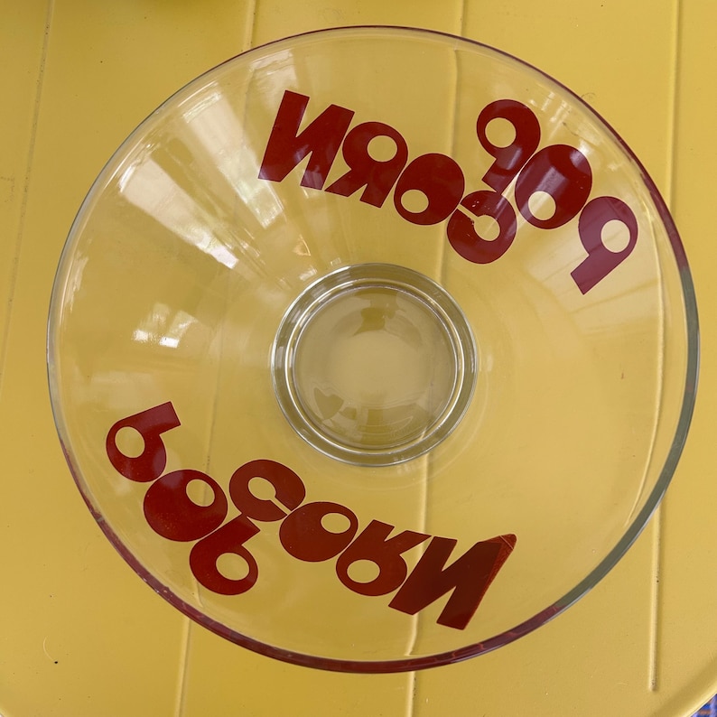 Op de afbeelding: Een heldere glazen kom met het woord POPCORN in dikke rode letters twee keer afgedrukt op het binnenoppervlak. De kom heeft een brede, ronde opening en een gladde, transparante afwerking, ontworpen voor het serveren van snacks.