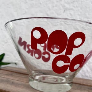 Op de afbeelding: Een heldere glazen kom met het woord POPCORN in dikke rode letters. De kom heeft een brede, uitlopende rand die taps toeloopt naar een ronde voet. De tekst is zichtbaar door het glas en toont de letters aan beide kanten van de kom.