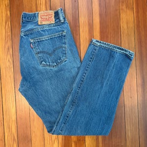 May include: Blue denim jeans featuring a classic five-pocket design, a button fly, and a tan leather patch on the back waistband with the text Levi Strauss & Co. 501 36 34. The jeans have a medium wash with visible stitching and a red tab.
