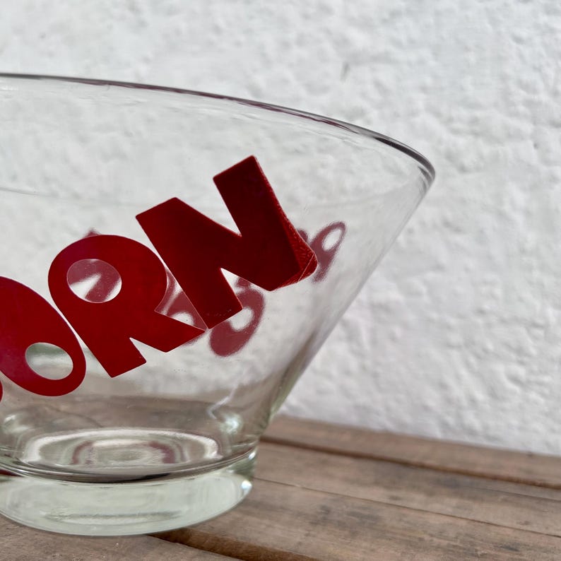 Op de afbeelding: Een heldere glazen kom met opvallende rode letters aan de buitenkant. De zichtbare tekst bevat de letters O, R en N, met extra gedeeltelijke rode tekens die door de transparante glazen wanden van de kom heen te zien zijn.