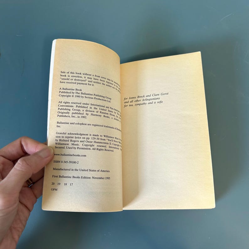 May include: An open book with text on the pages. The left page details publication and copyright information. The right page contains a dedication. The book is held open by a hand, against a blue background.