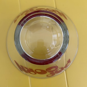 Op de afbeelding: Een heldere glazen kom met een dikke, donkerrode rand aan de bovenzijde. Het woord POPCORN is in grote, dikgedrukte, donkerrode letters op de buitenkant van de kom gedrukt, zichtbaar door het transparante glas vanuit dit bovenaanzicht.