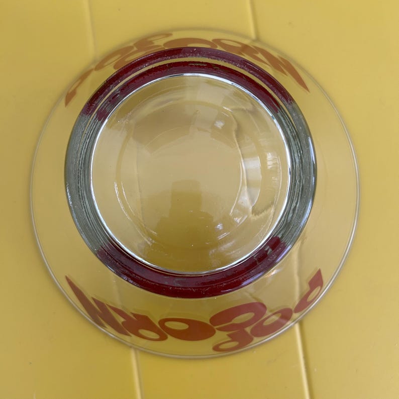 Op de afbeelding: Een heldere glazen kom met een dikke, donkerrode rand aan de bovenzijde. Het woord POPCORN is in grote, dikgedrukte, donkerrode letters op de buitenkant van de kom gedrukt, zichtbaar door het transparante glas vanuit dit bovenaanzicht.