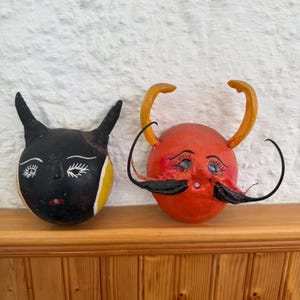 May include: Two round, hand-painted masks with horns. The left mask is black with yellow side accents, white painted eyes, and a small red mouth. The right mask is orange with yellow horns, long black painted eyelashes, and a prominent black mustache.