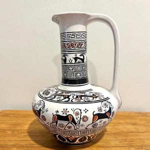 May include: A white ceramic pitcher featuring black and orange hand-painted motifs. The design includes geometric patterns, floral elements, and stylized animal figures resembling ibexes encircling the neck and the rounded body of the vessel.