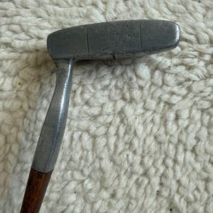 May include: A vintage golf putter featuring a weathered, dark grey metal head with a visible engraved letter O and a vertical alignment line. The putter includes a smooth, dark brown wooden shaft connected to the metal head by a silver-toned metal hosel.