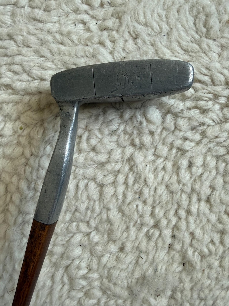 May include: A vintage golf putter featuring a weathered, dark grey metal head with a visible engraved letter O and a vertical alignment line. The putter includes a smooth, dark brown wooden shaft connected to the metal head by a silver-toned metal hosel.