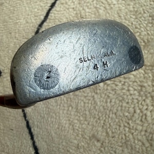 May include: A silver-toned metal golf putter head with a rounded shape and a textured surface. The face features the stamped text "SELMA, ALA." above "4 H" and a circular marking with the number "2" inside on the left side of the club face.