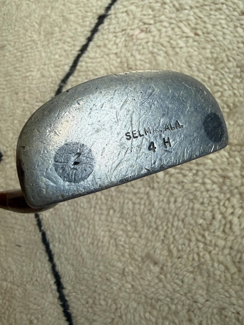 May include: A silver-toned metal golf putter head with a rounded shape and a textured surface. The face features the stamped text "SELMA, ALA." above "4 H" and a circular marking with the number "2" inside on the left side of the club face.