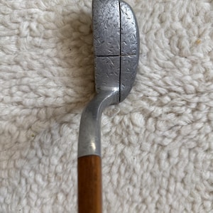 May include: A golf putter featuring a silver-toned metal head with a textured, hammered finish and a single vertical alignment line. The club head is attached to a smooth, cylindrical wooden shaft that extends downward from the metal hosel.