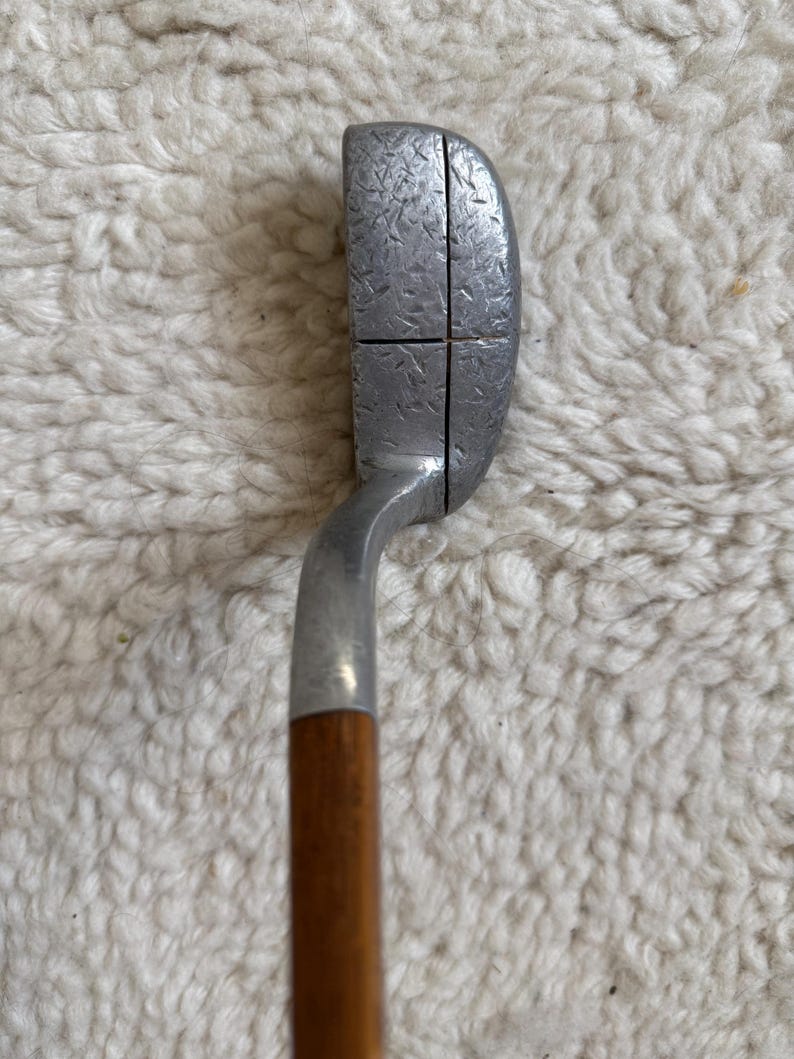 May include: A golf putter featuring a silver-toned metal head with a textured, hammered finish and a single vertical alignment line. The club head is attached to a smooth, cylindrical wooden shaft that extends downward from the metal hosel.