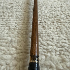 May include: A long, slender wooden shaft with a polished brown grain finish, tapering toward the top. The lower section features a black wrapped handle with a small gold-lettered label partially visible near the transition point of the wood.
