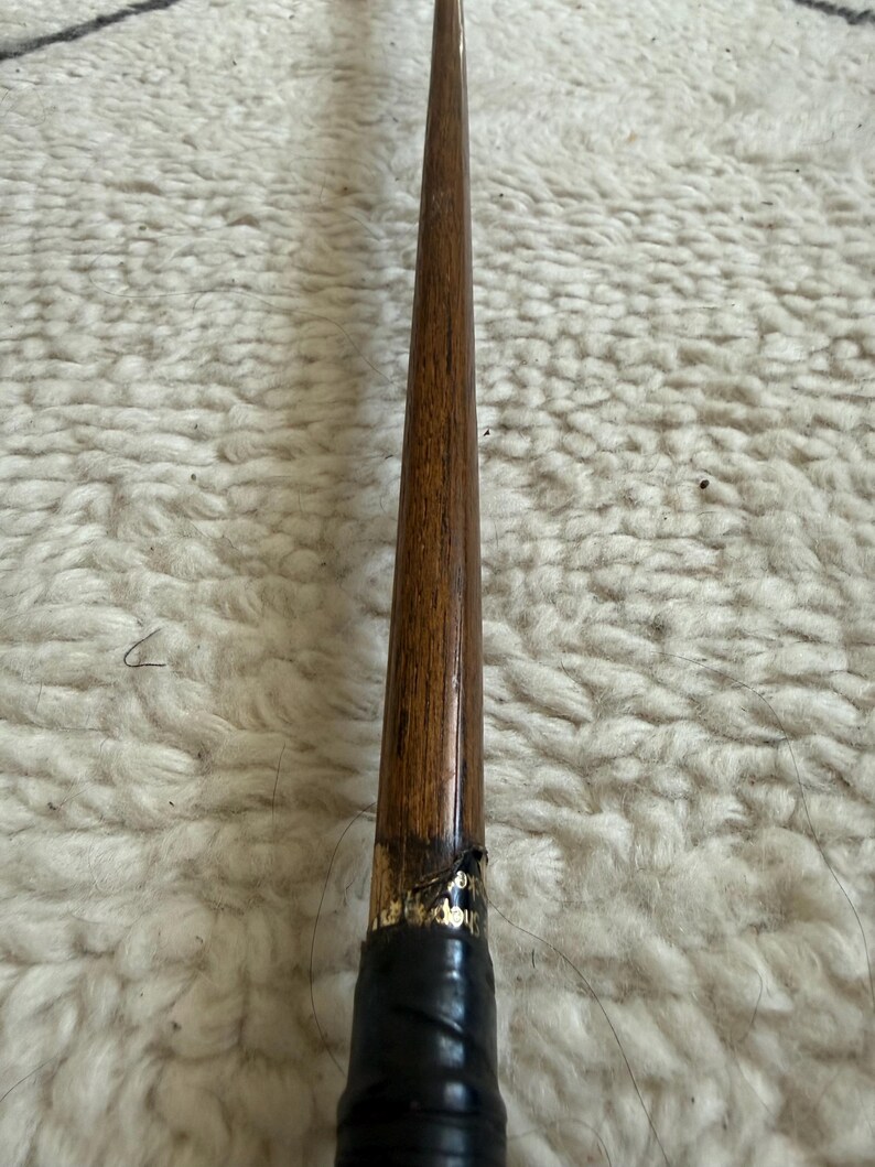 May include: A long, slender wooden shaft with a polished brown grain finish, tapering toward the top. The lower section features a black wrapped handle with a small gold-lettered label partially visible near the transition point of the wood.