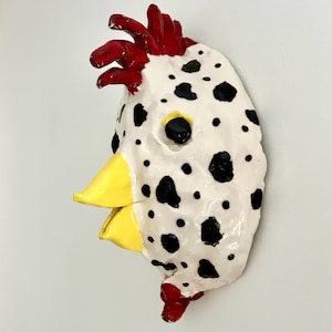 May include: A ceramic wall sculpture shaped like a chicken head, featuring a white base with black spots, a bright yellow beak, and a glossy red comb and wattle. The surface has a textured, hand-molded appearance with a shiny, reflective finish.