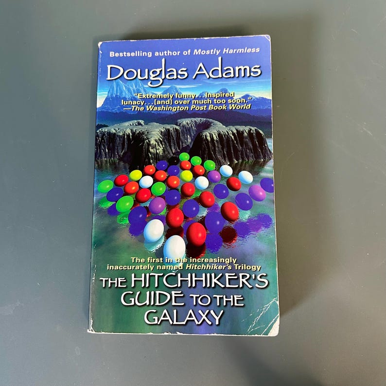 May include: A copy of "The Hitchhiker's Guide to the Galaxy" by Douglas Adams. The book cover displays a mountain scene and colorful spheres. The text includes quotes and mentions the Hitchhiker's Trilogy.