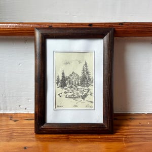May include: A framed black and white ink drawing on off-white paper depicting a mountain landscape with pine trees and rocky terrain. The artwork is enclosed in a dark brown wooden frame with a white mat. The text Kriváň 55/50 is visible.