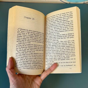 May include: An open book with text on yellowed pages. The left page shows "Chapter 24" and a large initial letter. The right page continues the text. The book is held open by a hand.