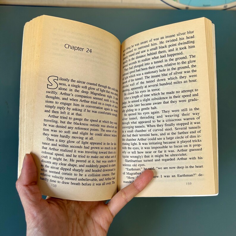 May include: An open book with text on yellowed pages. The left page shows "Chapter 24" and a large initial letter. The right page continues the text. The book is held open by a hand.