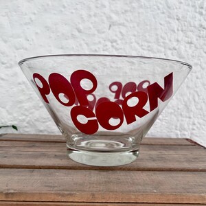 Op de afbeelding: Een heldere glazen kom met het woord POPCORN in dikke rode letters op de voorzijde. De kom heeft een brede, uitlopende rand die taps toeloopt naar een ronde voet, wat zorgt voor een eenvoudig en functioneel ontwerp voor het serveren.