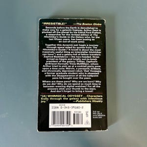 May include: A paperback book with a black cover and white text. The text includes the title "The Hitchhiker's Guide to the Galaxy" and a quote. The book has a barcode and the price.