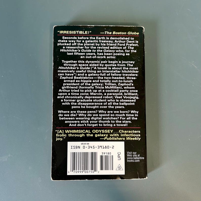 May include: A paperback book with a black cover and white text. The text includes the title "The Hitchhiker's Guide to the Galaxy" and a quote. The book has a barcode and the price.