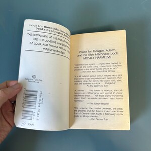 May include: An open book, "Mostly Harmless" by Douglas Adams, with text visible. The pages show reviews and quotes. The cover displays the title and author. The book is open on a blue surface.