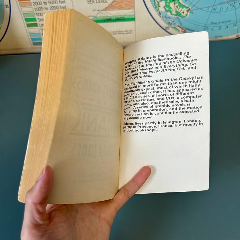 May include: A book with yellowed pages is held open, displaying printed text. The text details author Douglas Adams and his book "The Hitchhiker's Guide to the Galaxy". The book is set against a backdrop of a world map.