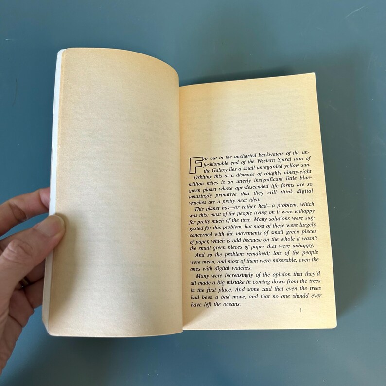 May include: An open book with yellowed pages. The text is in black, with a large initial letter at the start of the first paragraph. The book is held open, revealing the text and the edge of the pages.