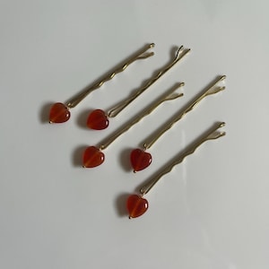 Pode incluir: Cinco grampos de cabelo dourados, cada um adornado com uma pedra preciosa em forma de coração, de cor vermelho-alaranjado. Os grampos estão dispostos diagonalmente sobre um fundo branco, apresentando um acessório de cabelo simples e elegante.