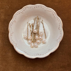 Pode incluir: Um prato de porcelana branca com borda festonada e design floral, contendo um conjunto de grampos de cabelo dourados com detalhes de pérolas.