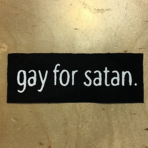 May include: A black rectangular patch with the white text "gay for satan." The patch is made of a woven material and is displayed against a light brown wooden surface.
