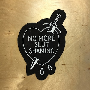 May include: Black fabric patch in the shape of a heart with a white dagger piercing through it. The heart has the text "NO MORE SLUT SHAMING" in white. Three white teardrop shapes are below the dagger.