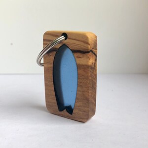 Può includere: Un portachiavi in legno con un design a forma di tavola da surf blu. Il portachiavi ha un anello in metallo argentato.